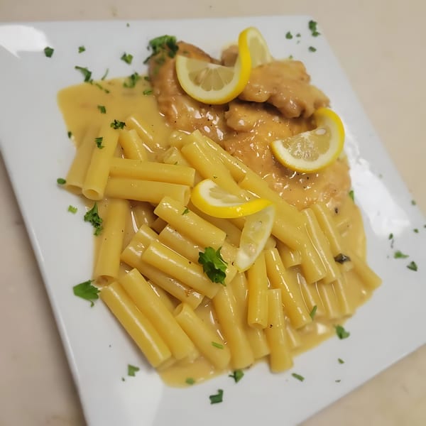 Midways De La Roma's chicken piccata with rigatoni pasta, lemon slices, and parsley on a white plate.