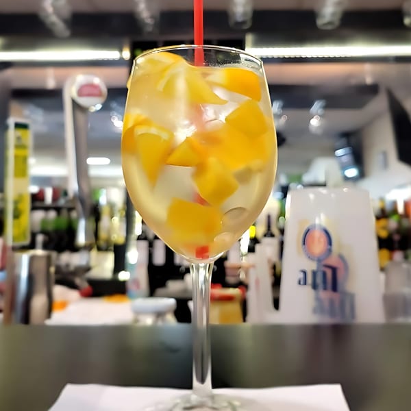 A glass of white sangria with fruit and a red straw on a bar.