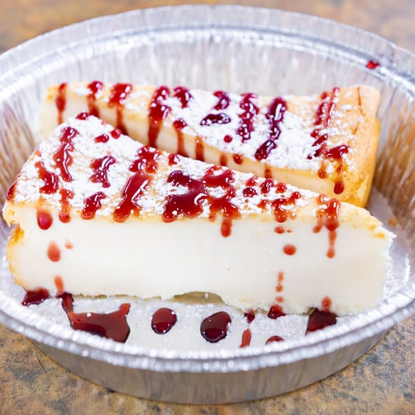 Two slices of cheesecake with powdered sugar and raspberry drizzle in a foil container.