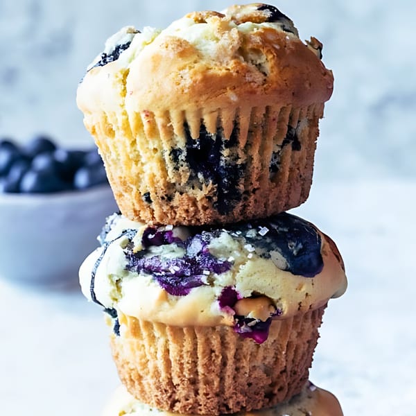 A stack of three blueberry muffins, with a bowl of blueberries blurred in the background.