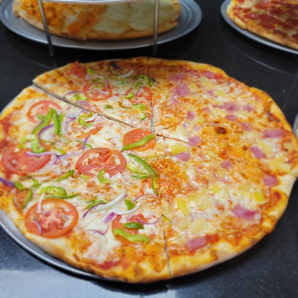 Two pizzas on metal trays, one topped with vegetables, the other with ham and pineapple.