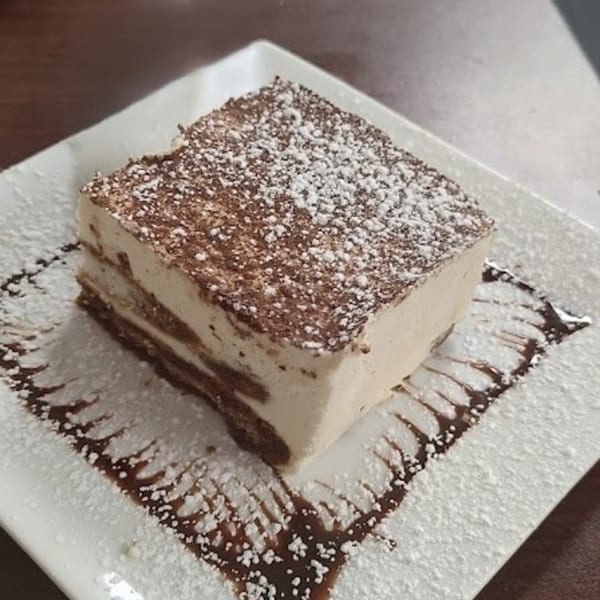 A square slice of tiramisu, dusted with powdered sugar and cocoa, on a white plate with chocolate drizzle.