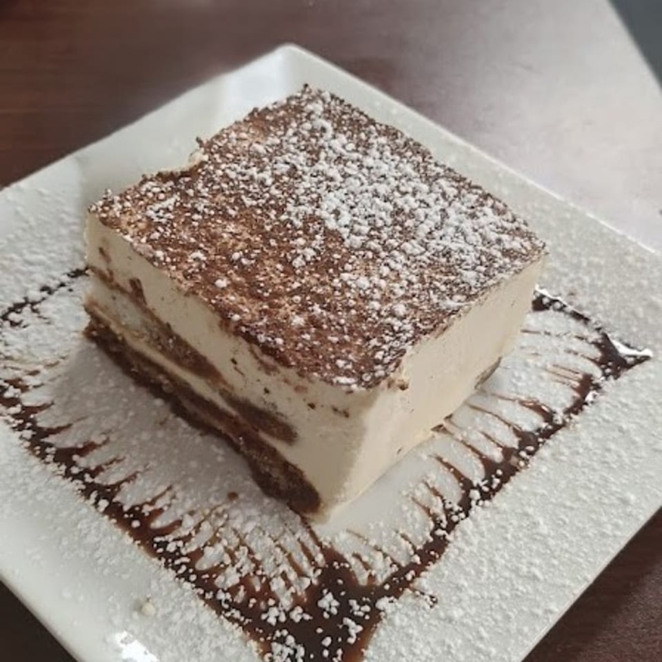 A square slice of tiramisu, dusted with powdered sugar and cocoa, on a white plate with chocolate drizzle.