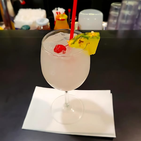 A white cocktail with ice, a red cherry, and a pineapple wedge, on a white napkin.